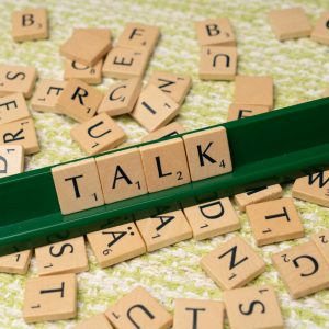 Scrabble tiles arranged to spell 'Talk' on a green tray, surrounded by scattered tiles on a textured surface.