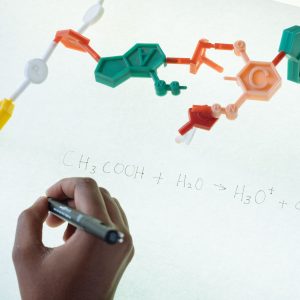 Anonymous chemist writing with marker on illuminated magnetic board formula of Hydronium and Acetate production after studying plastic model of molecule in laboratory
